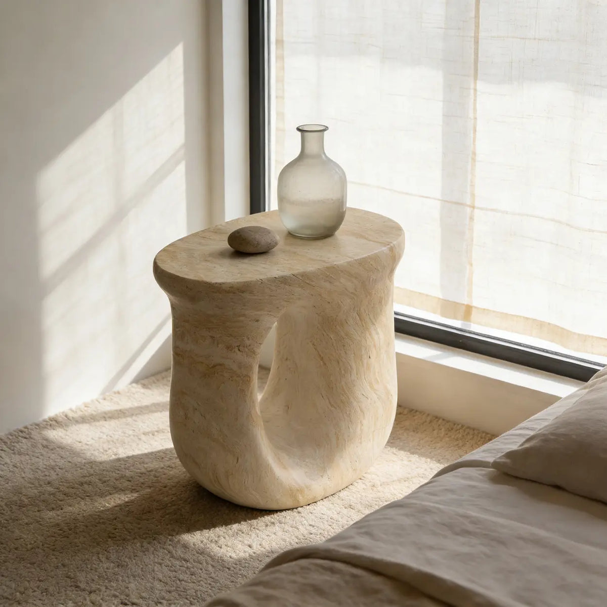 Faux travertine stone oval side table with a curved open base by a sunlit bedroom window, styled with glass decor