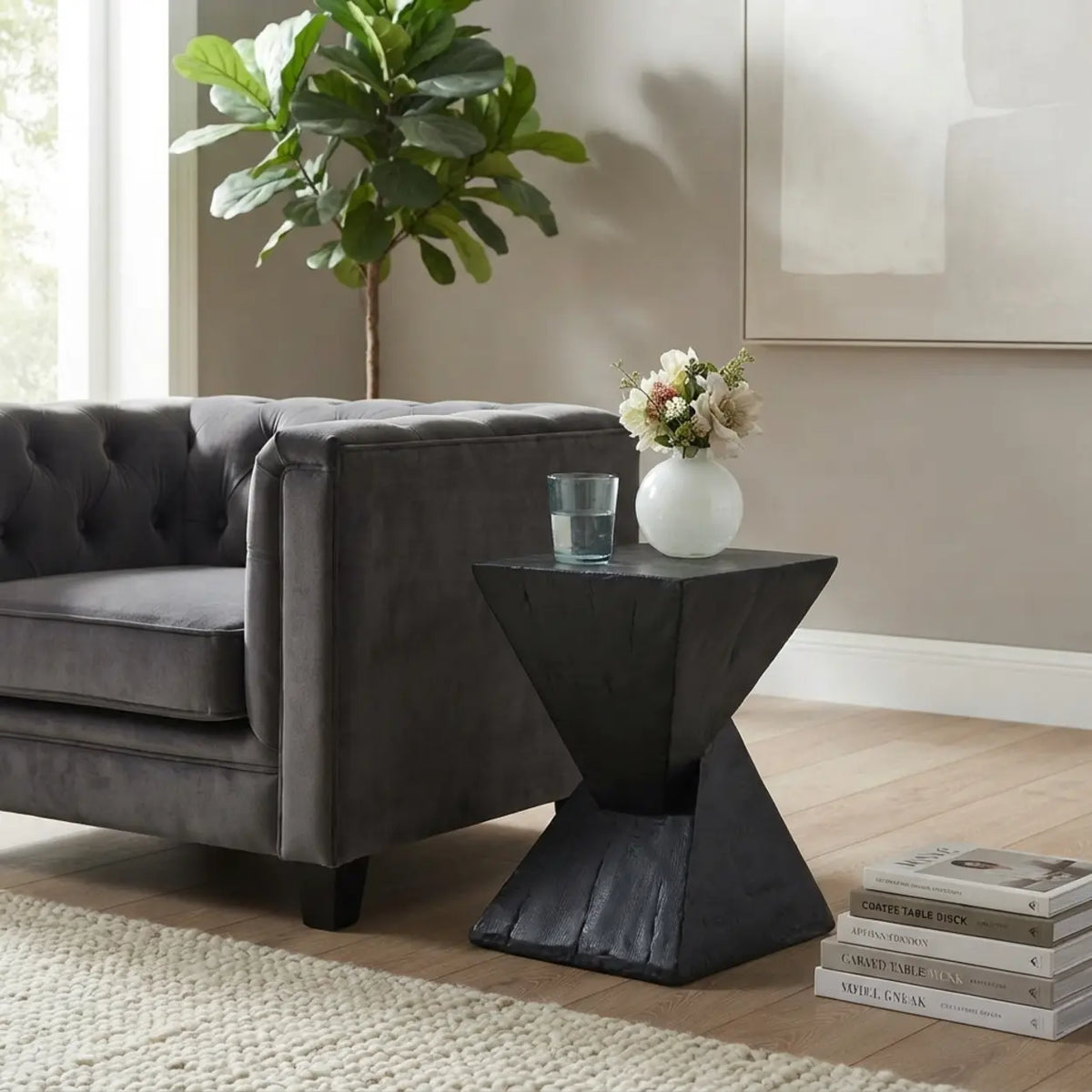 Black faux travertine stone side table with a twisted geometric pedestal beside a gray sofa, styled with a flower vase and glass