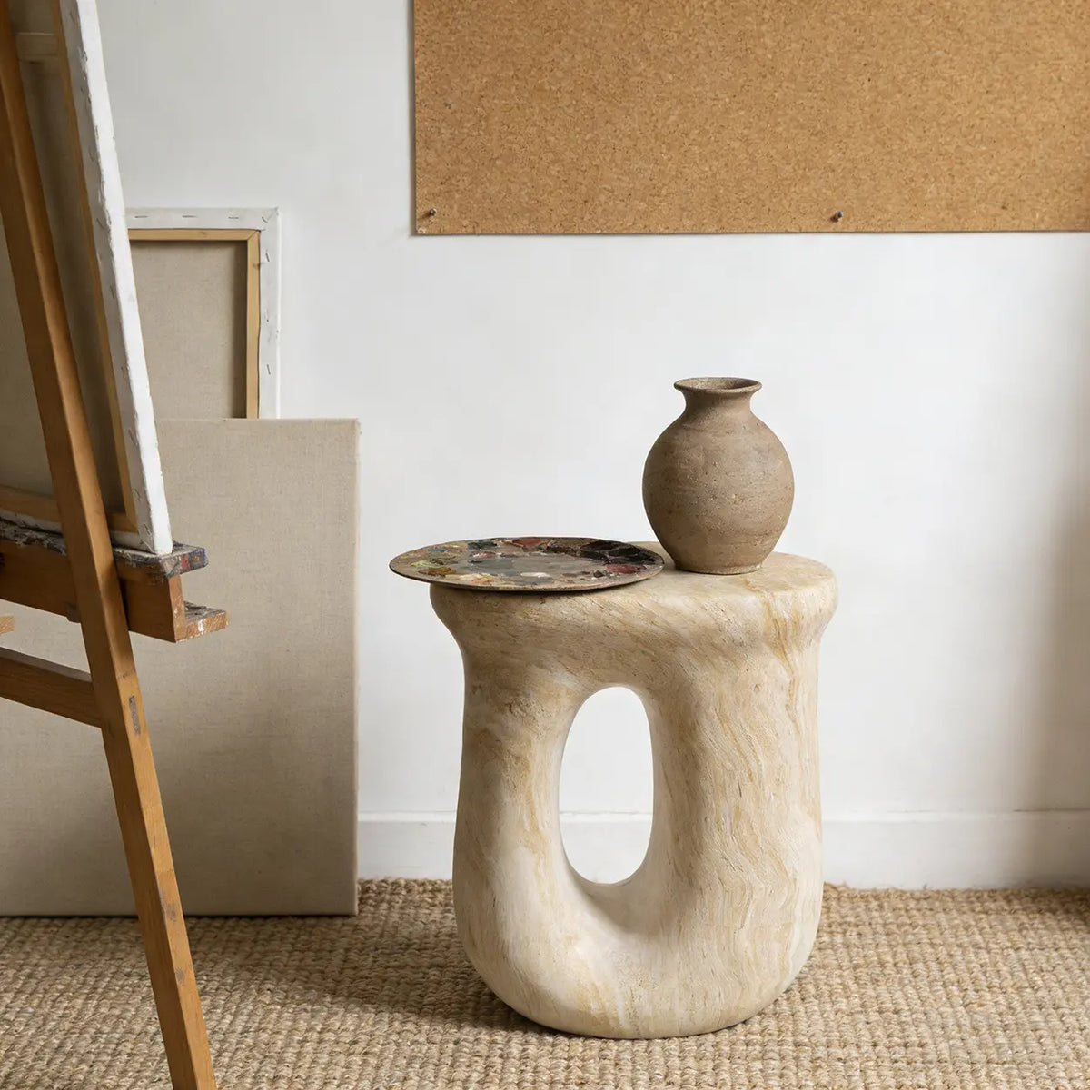 Faux travertine stone oval side table with a sculptural curved base holding a vase and artist palette beside an easel
