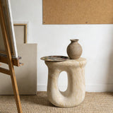 Faux travertine stone oval side table with a sculptural curved base holding a vase and artist palette beside an easel