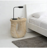 Faux travertine stone oval side table with a curved open base beside a bed, styled with a black table lamp and bowl