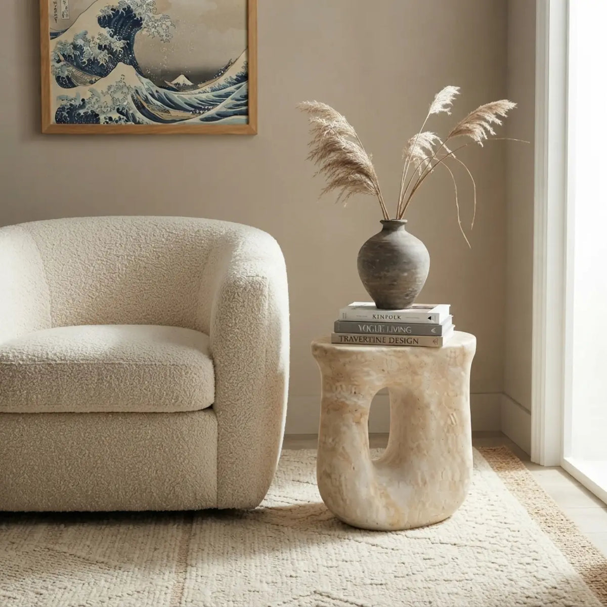 Faux travertine stone oval side table with a curved open base beside a boucle chair, topped with books and a decorative vase