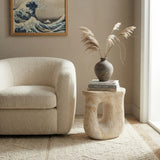 Faux travertine stone oval side table with a curved open base beside a boucle chair, topped with books and a decorative vase