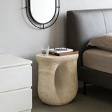 Faux travertine stone oval bedside table with a rounded hollow base beside a bed, holding a glass and book tray