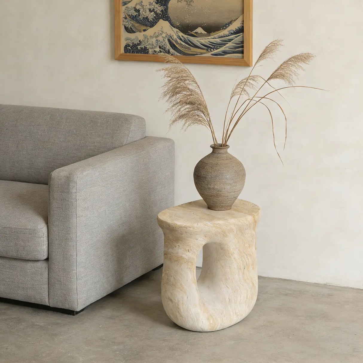 Faux travertine stone oval accent table with a curved open base beside a gray sofa, styled with a vase