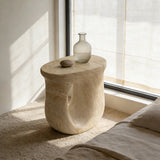 Faux travertine stone oval side table with a curved open base by a sunlit bedroom window, styled with glass decor