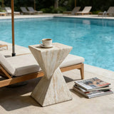 Travertine faux stone side table with a twisted geometric pedestal beside a pool lounge chair, topped with a cup and magazines