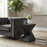 Black faux travertine stone side table with a twisted geometric pedestal beside a gray sofa, styled with a flower vase and glass