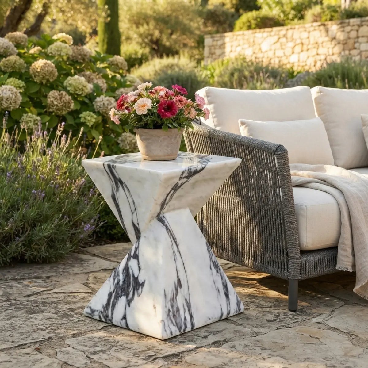 White faux stone side table with dark veining and a twisted geometric pedestal beside an outdoor lounge chair with flowers