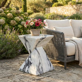 White faux stone side table with dark veining and a twisted geometric pedestal beside an outdoor lounge chair with flowers